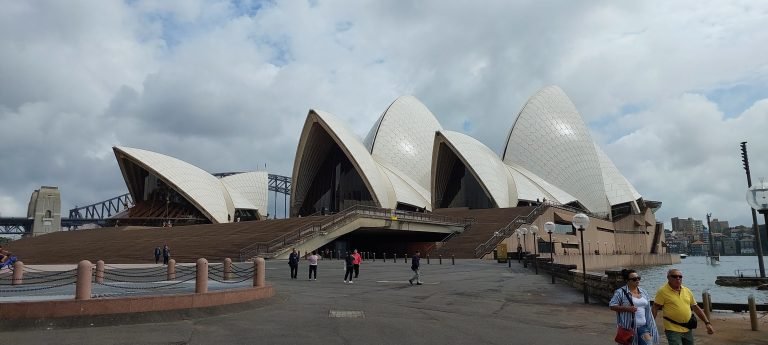 Close-up view of the Sydney Opera House sails with people standing on the forecourt under a cloudy sky.