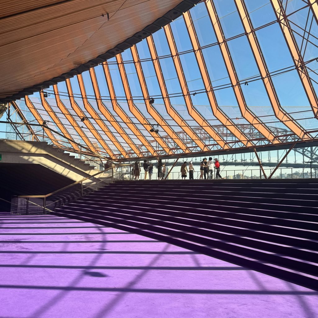 Modern interior walkway in the Sydney Opera House with a glass ceiling, purple carpeted steps, and visitors overlooking Sydney Harbour.