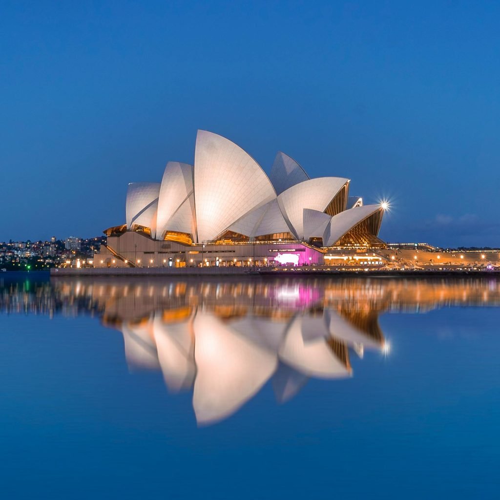 Sydney Opera House tours tickets: Sydney Opera House at night reflected in calm harbour water, with the sails glowing white against a deep blue sky.