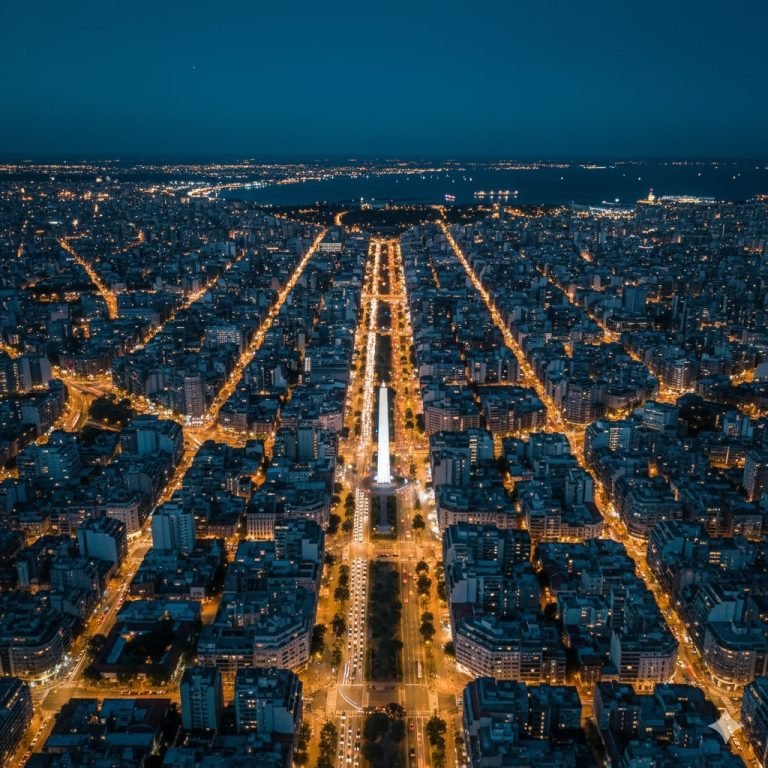 Aerial night view of Buenos Aires with the illuminated Obelisk along Avenida 9 de Julio and city lights stretching toward the Río de la Plata.