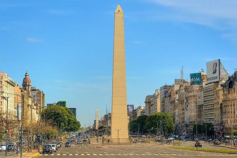 The Obelisk of Buenos Aires standing along Avenida 9 de Julio on a clear sunny day, with traffic and historic buildings lining the wide boulevard.