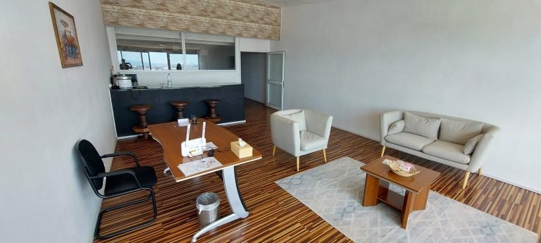 Bright, minimalist hotel suite living area with a sofa, armchair, desk, and a bar-style kitchenette in the background.