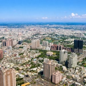 A bright aerial view of Kaohsiung, Taiwan, with the city skyline, green parks, and modern buildings under clear blue skies.