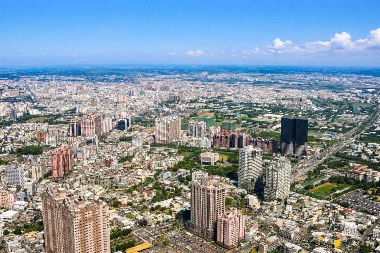 A bright aerial view of Kaohsiung, Taiwan, with the city skyline, green parks, and modern buildings under clear blue skies.