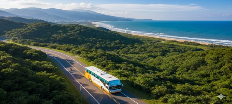An aerial view of a modern white and blue coach bus traveling along a winding coastal highway in the Taiwan countryside between Kaohsiung and Kenting, with the turquoise ocean on one side and lush green hills on the other.