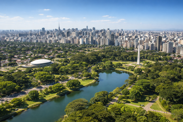Aerial view of São Paulo on a sunny day, showing Ibirapuera Park surrounded by high-rise buildings and clear blue skies.
