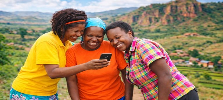Malagasy women smiling together while looking at a smartphone in rural Madagascar, with green hills and red highlands in the background.