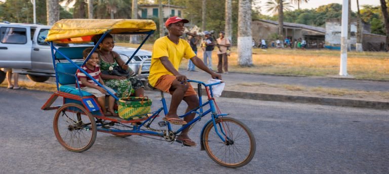 Cyclo-pousse in Madagascar with smiling passengers, a child and an adult seated behind the driver as the bicycle taxi moves through a palm-lined town.