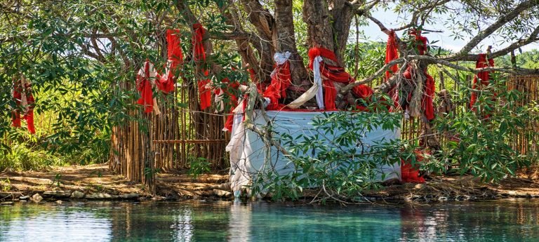 Sacred fady shrine at a sacred lake in Majunga (Mahajanga), Madagascar, with red and white ceremonial cloths tied to a tree above calm turquoise water.