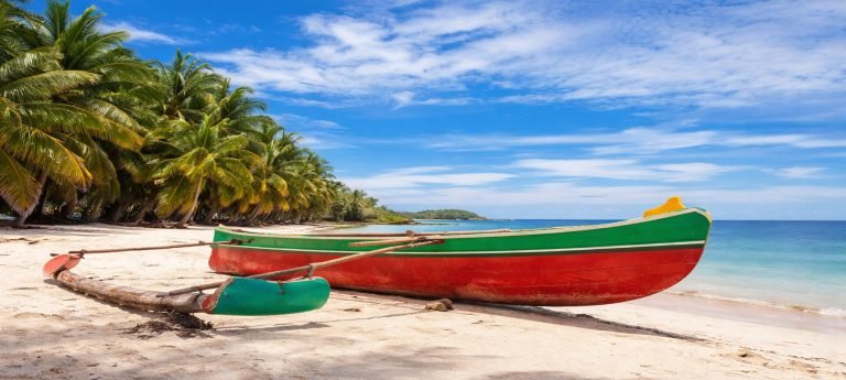 Wooden fishing boat resting on white sand beside a calm turquoise ocean, with palm trees lining the beach under a bright blue sky.