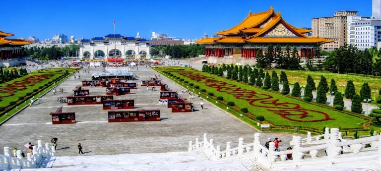 Things to do in Taipei: Wide, sunlit view of Chiang Kai-shek Memorial Hall Square in Taipei, with ornate buildings, manicured gardens, and visitors under a bright blue sky.