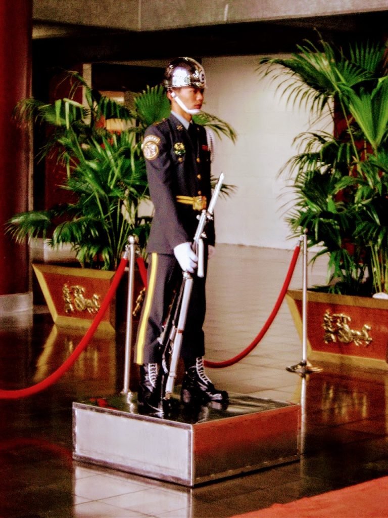 Ceremonial guard in Taiwan wearing a formal uniform and silver helmet, standing at attention with a rifle indoors.