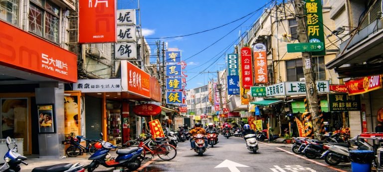 Sunlit street scene in Taiwan with scooters parked along both sides, riders driving down a narrow road, and colorful Chinese shop signs under a bright blue sky.