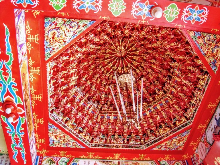 Intricately decorated temple ceiling in Taiwan with red wood panels, gold carvings, and colorful traditional patterns.