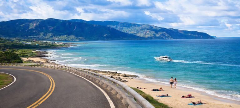 Scenic coastal road in Kenting, Taiwan overlooking turquoise ocean, sandy beach, and green mountains with people relaxing on the shore and a boat offshore.