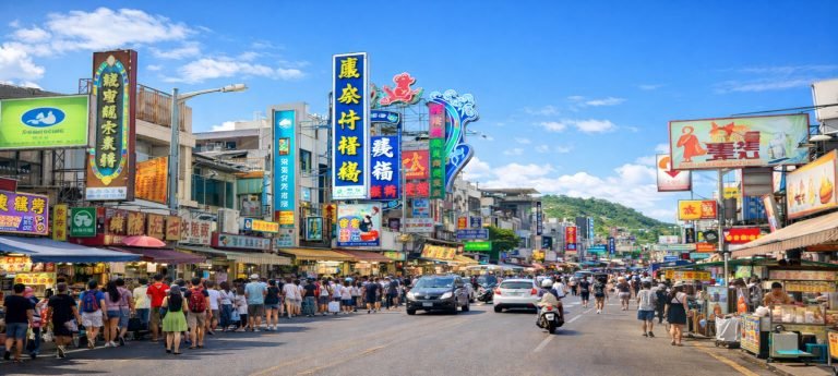 Kenting Taiwan: Daytime street market in Kenting, Taiwan with crowds, food stalls, and colorful shop signs under a bright blue sky.