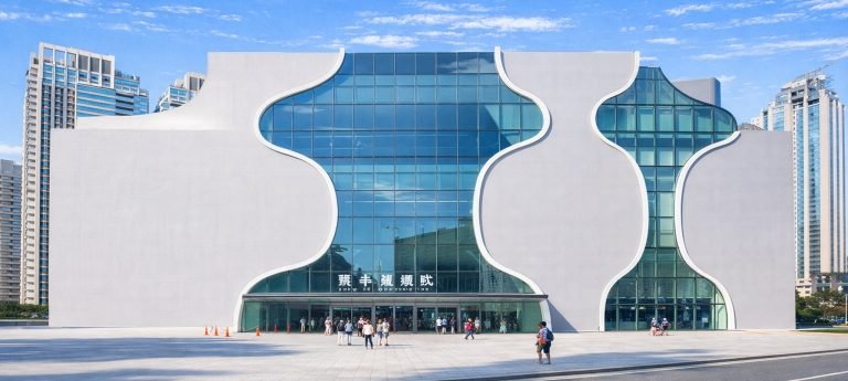 The modern facade of the National Taichung Theater in Taiwan, featuring its iconic curved glass windows and white "sound cave" architecture under a clear blue sky.