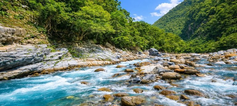 Sunlit turquoise river flowing through Taroko Gorge with marble rocks, forested canyon walls, and blue skies above