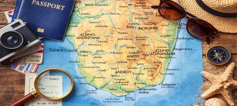 Flat lay travel planning scene with a map of Southern Madagascar showing Toliara and Fort Dauphin, surrounded by a passport, sunglasses, camera, compass, and seashells.
