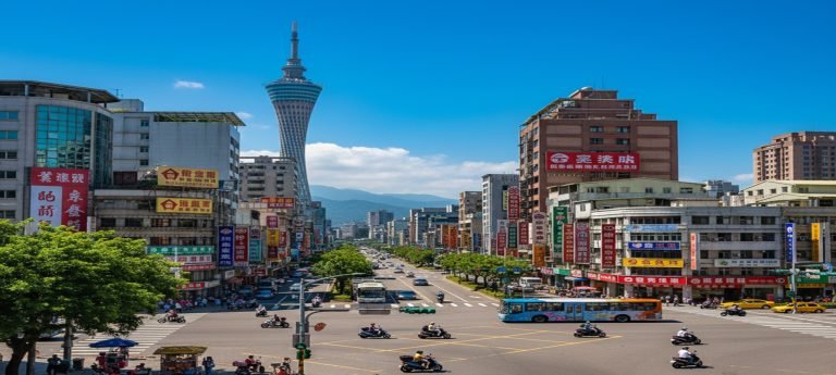 A wide, sunlit city intersection in Kaohsiung, Taiwan, filled with scooters and a blue bus, with colorful building signage and a tall landmark tower visible in the background under a blue sky.