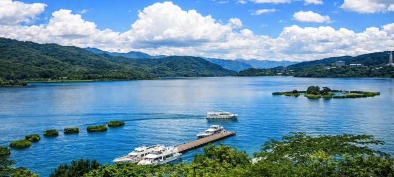 Sun Moon Lake pier with sightseeing boats and small islands, surrounded by green mountains under blue skies in Taiwan.