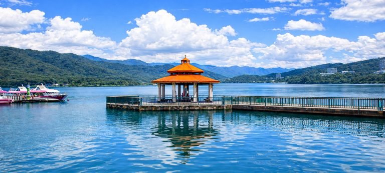 Orange-roof pavilion pier at Sun Moon Lake with boats, calm water reflections, and green mountains under blue skies.