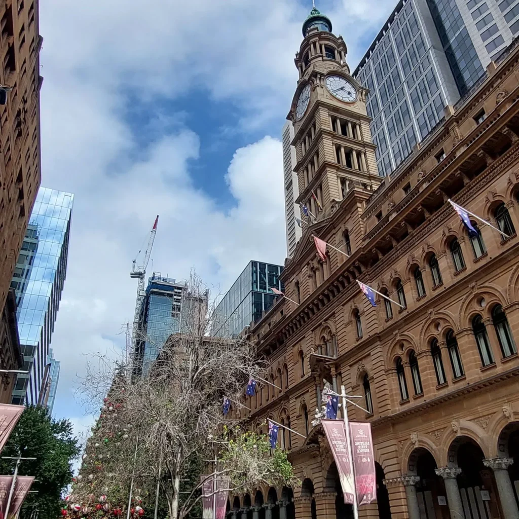 Historic sandstone building with a tall clock tower in Sydney CBD, framed by modern skyscrapers and a partly cloudy blue sky.