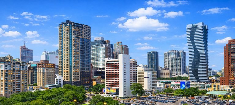 A view of the Taichung city skyline on a clear day with blue skies and scattered clouds, featuring modern high-rise buildings, including a light yellow residential tower on the left, a white building with red accents in the center, and a uniquely designed wavy-structure tower on the right, with a large foreground parking lot filled with vehicles.