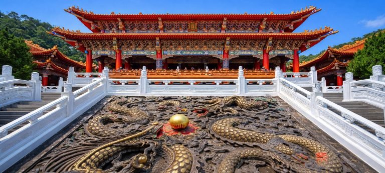 Ornate Taiwanese temple in Taichung with colorful red-and-gold roof and large dragon relief sculpture on the stairway under a bright blue sky.