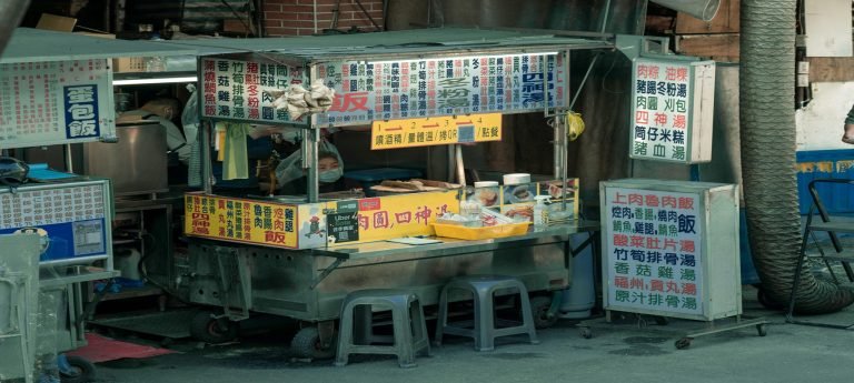 Traditional Taiwanese street food stall in Taipei with Chinese menu boards, cooking pots, stools, and a vendor preparing food.