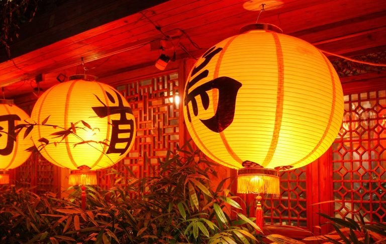 Traditional Taiwanese lanterns glowing in warm orange light, hanging under a wooden roof beside bamboo plants and red lattice windows.