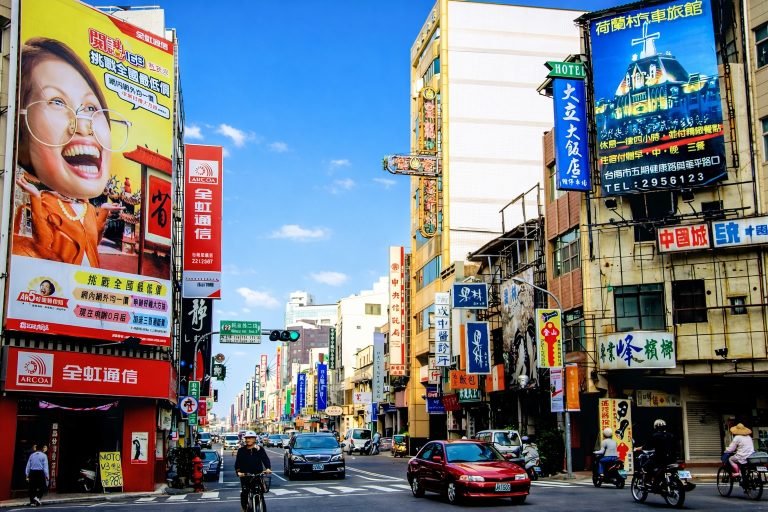 A sunny street scene in Taiwan with blue skies, colorful Chinese shop signs, a large billboard, cars, scooters, and cyclists at a busy intersection.