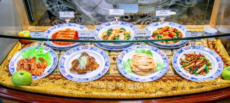 Things to do in Taiwan: Taiwanese food court display case featuring multiple plated dishes on blue-and-white porcelain plates, arranged behind glass with small menu labels.