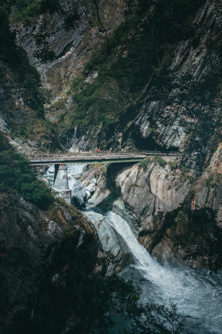 A narrow bridge crossing Taroko Gorge in Taiwan, suspended above a rushing turquoise river cutting through steep marble cliffs and dense forest.