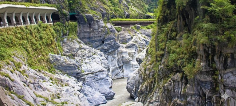 A high-resolution landscape view of Taroko Gorge in Taiwan, featuring steep, light-gray marble cliffs covered in lush green vegetation under bright, sunny afternoon light. A white architectural viewing gallery with supportive pillars is built into the cliffside overlooking a narrow river winding through the canyon floor.
