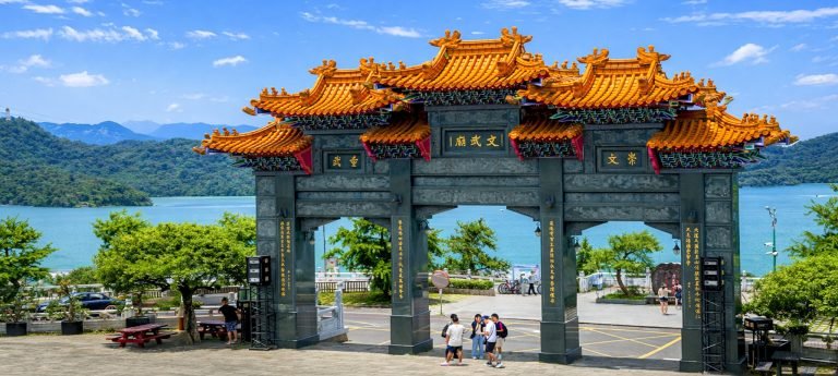 The ornate stone Paifang gate of Wenwu Temple at Sun Moon Lake, Taiwan, featuring traditional orange tiled roofs and intricate carvings with the turquoise lake and mountains in the background.