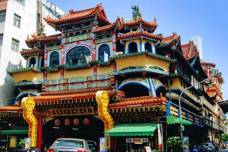 Crisp, sunny-day photo of a colorful multi-level Taiwanese temple with ornate red-and-gold rooflines and intricate carvings, surrounded by city buildings.