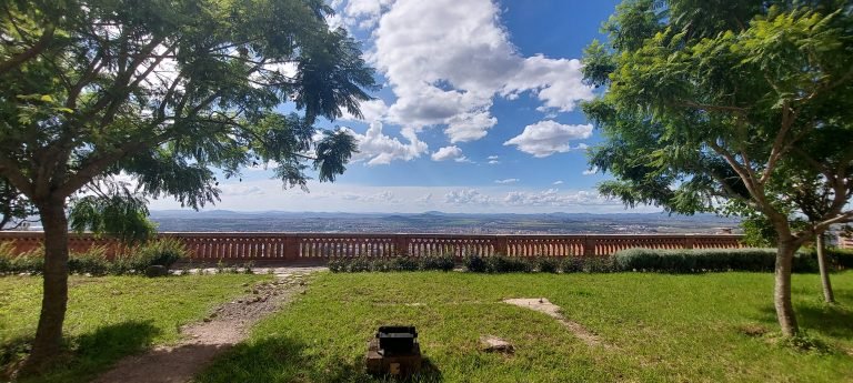 Lawns and viewpoint outside the Queen’s Palace (Rova) in Antananarivo, framed by trees and overlooking the city of Antananarivo down below under a bright blue sky