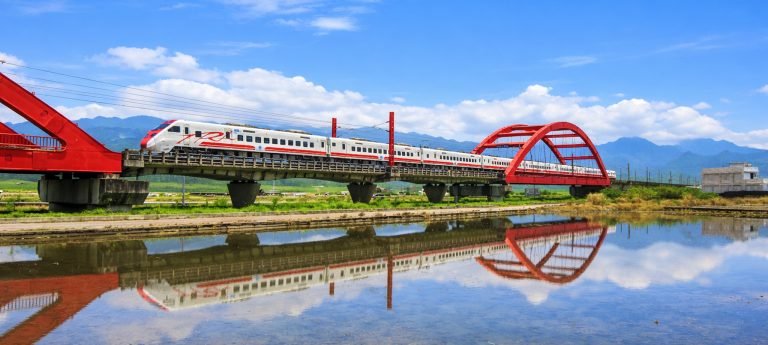High-speed train crossing a red railway bridge with blue skies and mountain reflections in rural Taiwan