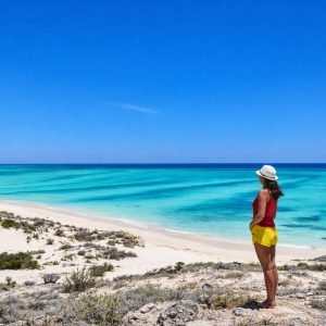 Woman standing on coastal dunes overlooking a bright turquoise sea and white-sand beach under a clear blue sky.