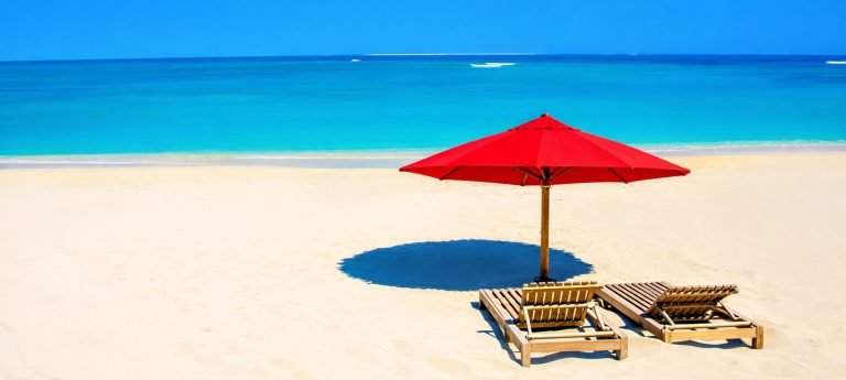 Two wooden lounge chairs under a bright red beach umbrella on soft white sand, facing a calm turquoise ocean under a clear blue sky in southern madagascar