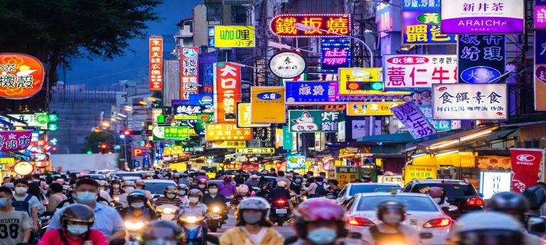 Bustling night market street in Taichung, Taiwan with neon signs, scooters, and crowds at dusk.