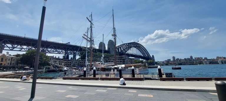 View of Sydney Harbour with the Sydney Harbour Bridge, waterfront promenade, and historic ships docked near Circular Quay.
