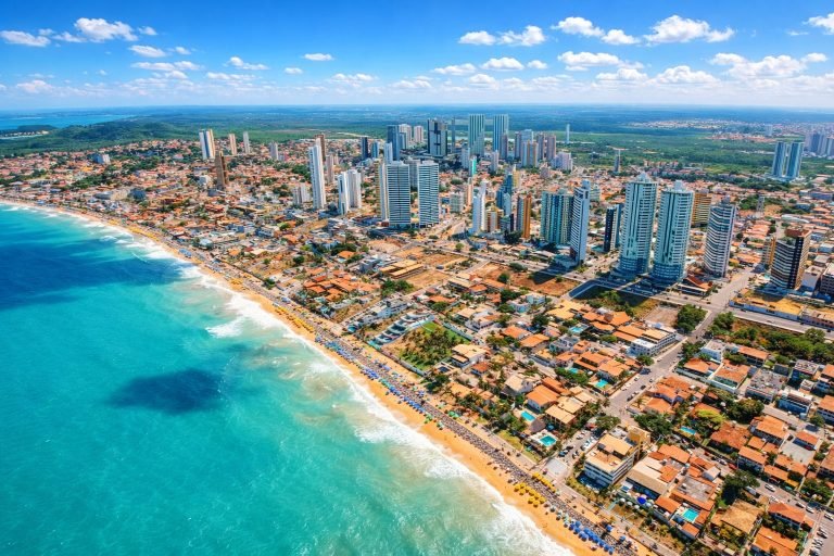 Aerial view of Salvador Brazil coastline with turquoise ocean, golden beach, and modern high-rise skyline under bright blue skies.