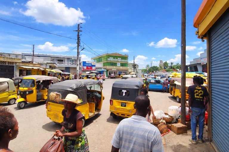 Busy street scene in Madagascar with yellow bajaj tuk-tuks, pedestrians, and market stalls under bright blue skies.