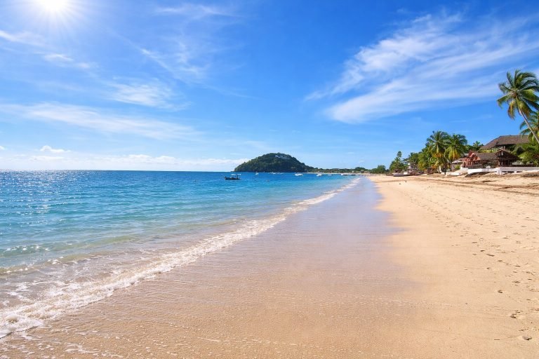 Getting Around Madagascar: Bright sandy beach on Nosy Be with turquoise water, palm trees, and clear blue skies on a sunny day.