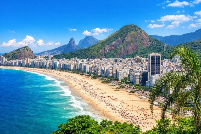 Bright sunny view of Copacabana Beach in Rio de Janeiro with turquoise ocean, golden sand, and mountains under clear blue skies. Caption Description