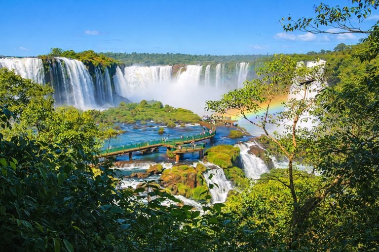 Iguazu Falls Brazil panoramic view with boardwalk platform, rainbow mist, and lush rainforest under bright blue skies Caption Description