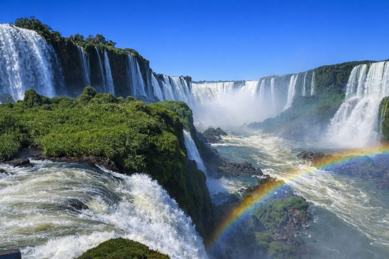 Brazil Itinerary: Iguazu Falls from the Brazilian side with powerful cascading waterfalls, lush green cliffs, mist rising into a clear blue sky, and a rainbow forming over the river below.