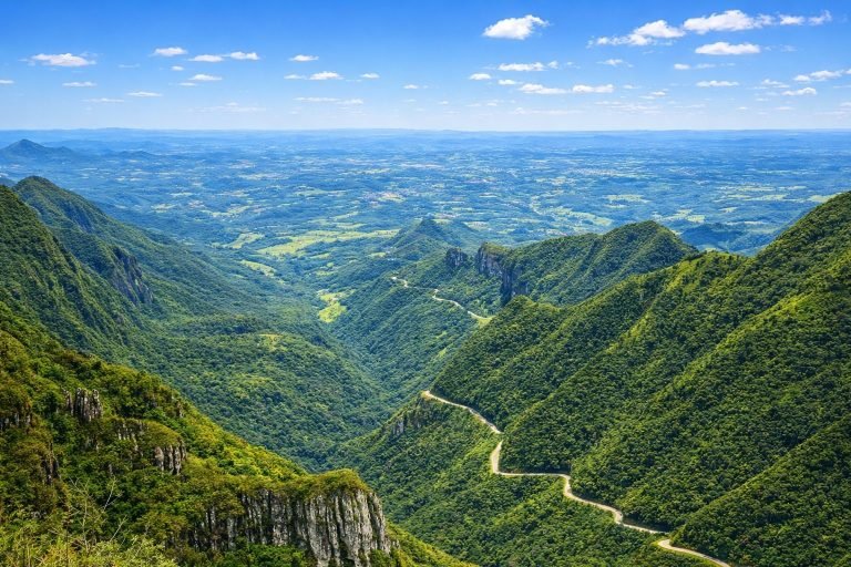 Panoramic view of lush green mountains in southern Brazil with a winding road cutting through the valley under a bright blue sky.